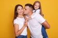 Horizontal shot of happy young family. Husband kissing wife and piggybacking thier little charming daughter, cute girl being close