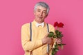 Horizontal shot of impressed grey haired elderly man in formal fashionable clothes, keeps hand near heart, holds red roses, makes