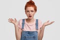 Horizontal shot of indignant puzzled red haired teenage girl shrugs shoulders with puzzlement, opens mouth, has Royalty Free Stock Photo