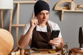 Horizontal shot of inspired excited man joiner wearing apron and cap sitting among his professional equipments and using phone,