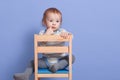 Horizontal shot of little boy sitting on stool, isolated on blue studio background, looks thoughtfuly aside, keeps finger near lip Royalty Free Stock Photo