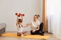 Horizontal shot of little girl raised arms with red dumbbells in hands, mother teaching her daughter doing fitness exercises at Royalty Free Stock Photo