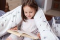 Horizontal shot of little girl reading under blanket with dandelion, having happy facial expression, looks exited, charming female Royalty Free Stock Photo