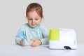 Horizontal shot of little girl sits at white desk with nebulous machine for health care, holds mask for making inhalation or Royalty Free Stock Photo