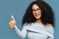 Horizontal shot of lovely dark skinned woman gives thumb up, wears fashionable sweater, eyewear, demonstrates approval, isolated