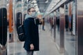 Horizontal shot of man traveler poses on metro station, wears medical mask during coronavirus time, waits for train, commutes to