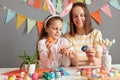 Horizontal shot of optimistic cheerful mother and her daughter painting Easter eggs, preparing for Easter, sitting at table with