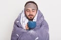 Horizontal shot of peaceful calm sleepy glad young man with beard, covering himself with blanket, closing eyes, holding cup with