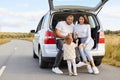 Horizontal shot of positive delighted family with infant kid drinking hot tea from thermos sitting in car trunk, stop their Royalty Free Stock Photo