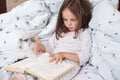Horizontal shot of pretty little girl reading interesting book in her room while lying in bed under blanket with dandelion, looks Royalty Free Stock Photo