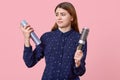 Horizontal shot of puzzled female model holds hairspray and comb, looks with indignation, dressed in blue shirt, poses over pink