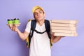 Horizontal shot of puzzled man in yellow cap and t-shirt, holding pizza and paper cups of coffee isolated on lilac background, Royalty Free Stock Photo
