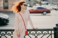 Horizontal shot of red haired woman wears sunglasses, focused aside, poses near hence at street, poses against road with transport