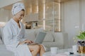 Horizontal shot of satisfied beautiful woman dressed in white soft bathrobe works on laptop computer surfes internet poses near Royalty Free Stock Photo