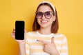 Horizontal shot of satisfied young woman wearing striped t-shirt posing isolated over yellow background, pointing at smart phone Royalty Free Stock Photo