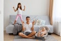 Horizontal shot of sdepressed attractive young adult man sitting on floor in lotus pose near sofa and looking after his little Royalty Free Stock Photo