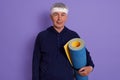 Horizontal shot of senior white haired male with head band and holding blue yoga mat in hands, posing isolated over lilac Royalty Free Stock Photo