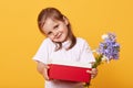 Horizontal shot of shy sweet lovely little girl smiling sincerely, looking directly at camera, holding red and white box, blue Royalty Free Stock Photo