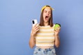 Horizontal shot of sleepy tired teen girl wearing striped T-shirt and baseball cap standing isolated over blue background, holding Royalty Free Stock Photo