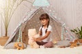 Horizontal shot of smiling satisfied little girl wearing white t shirt sitting on floor in wigwam and playing with teddy bear, Royalty Free Stock Photo