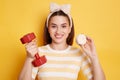 Horizontal shot of smiling satisfied young woman wearing striped shirt and hair band holding red dumbbell and marshmallow, posing Royalty Free Stock Photo