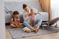 Horizontal shot of smiling woman sitting on floor near cough and playing with her children, enjoying her time together, mommy Royalty Free Stock Photo