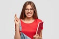 Horizontal shot of wishful Caucasian woman crosses fingers, dressed in casual clothes, holds red textbook, clenches