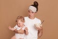 Horizontal shot of woman wearing white t shirt and sleeping mask, mother holding crying baby daughter and plate with kid`s food i Royalty Free Stock Photo