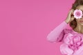 Horizontal shot of young curly haired woman holds pink gerbera daisy flower, covers eyes, dressed in fashionable pink dress, makes Royalty Free Stock Photo