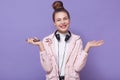 Horizontal shot of young pretty girl with hairbun listening to music and spreading palms aside, having headphones around neck,