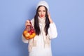 Horizontal shot of young woman holding shopping bag full of apples and oranges, dresses white warm clothing, standing against blue Royalty Free Stock Photo