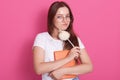 Horizontal shot of young woman with staright hair holding notebook and pen while standing against pink studio wall, looking Royalty Free Stock Photo