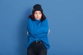 Horizontal studio picture of emotional upset female standing isolated over blue background, looking aside, hugging herself,