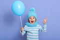 Horizontal studio picture of joyful sweet emotional little girl opening her mouth and mouth widely, raising arms, holding blue