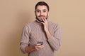 Horizontal studio picture of pleasant delighted young man looking directly at camera, touching chin with hand, holding his Royalty Free Stock Photo