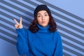 Horizontal studio shot of funny playful young attractive lady having stripes aback, raising hand, showing sign peace, wearing blue Royalty Free Stock Photo