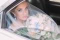 Horizontal view of the bride under the veil holding the wedding bouquet while sitting in the car.