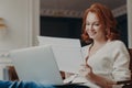 Horizontal view of pleased adult woman entrepreneur works from home, works remotely from home, looks through paper documents, uses Royalty Free Stock Photo