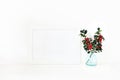 Horizontal white blank wooden frame mockup. Holly berry branches in blue glass vase on white table. Styled stock