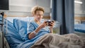 Hospital Ward: Handsome Young Boy Resting in Bed, Uses Smartphone, Plays Online Video Games on Int Royalty Free Stock Photo