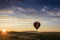 Hot Air Balloon in colorful rainbow stripes begins ascent over farm field as sun risesblue cloudy sky Royalty Free Stock Photo