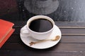 A hot cup of coffee is placed on a golden saucer and spoon. With a red book on a vintage wooden window sill or a window table with Royalty Free Stock Photo