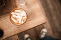 A hot cup of latte art coffee on a wooden table where people stand wearing blue sneakers on the floor at the coffee shop Royalty Free Stock Photo