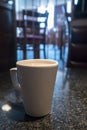 Hot drink in a white cup mug on a dark marble surface with blue toned blurred cafe restaurant scene in background.