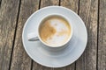 Hot foaming cappuccino in a white ceramich cup on a saucer stands on a wooden table, top view