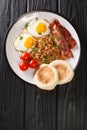 Hot hearty breakfast of fried eggs, beans, bacon, tomatoes and muffins close-up in a plate. vertical top view Royalty Free Stock Photo