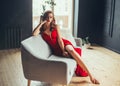 Hot young woman, dressed in a long scarlet red long dress, sexually shows her bare legs sitting on a grey sofa in a loft Royalty Free Stock Photo