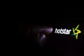 Hotstar streaming service logo on the screen in a dark room and a hand pointing at it. Hotstar is an Indian streaming service