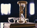 Hourglass, old book and eyeglasses on wooden table, dark background. Time flow concept. Wooden hourglass counting time