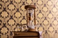 Hourglass, old book and eyeglasses on wooden table, pattern background. Sand falling down inside of hourglass. Hourglass